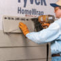 Construction worker holding up a piece of fiber cement siding board with a Malco FCG2 tool and using a staple gun to attach it to the side of a house