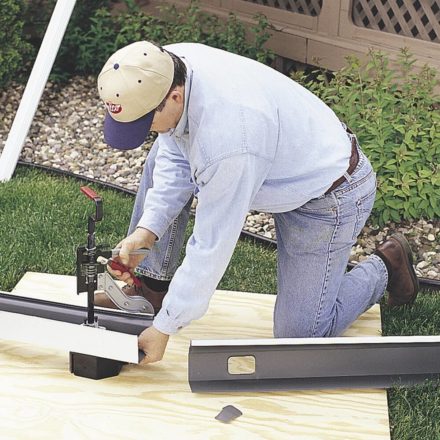 Construction worker using a Malco gutter outlet punch on a long section of white downspout